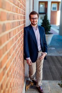 Picture of Chris Verrill in a Navy Jacket, Pink shirt, and tan pants leaning against a brick wall